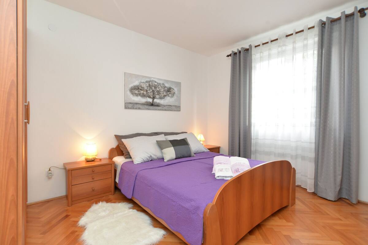 Une chambre à coucher avec un lit en bois, une couvre-lit violette, un tapis blanc et moelleux, une table de chevet avec une lampe et un tableau représentant un arbre accroché au mur.