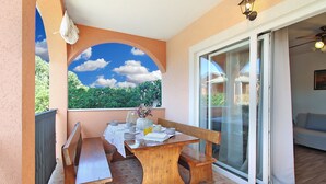 Varanda com mesa de madeira posta para dois, um banco e vista para o verde através de uma janela.