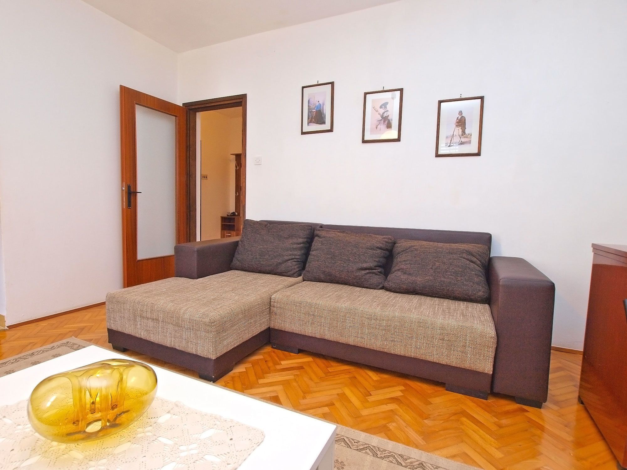 A living room with a sectional sofa, a wooden herringbone floor, and three framed pictures on the wall.