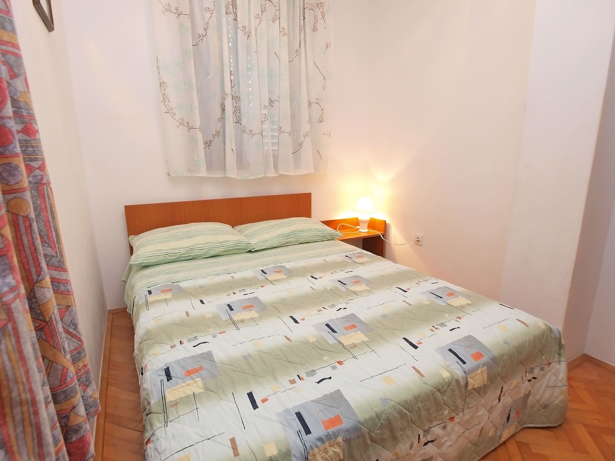 Una camera da letto con un letto, un comodino con una lampada e una finestra con tende leggere.
