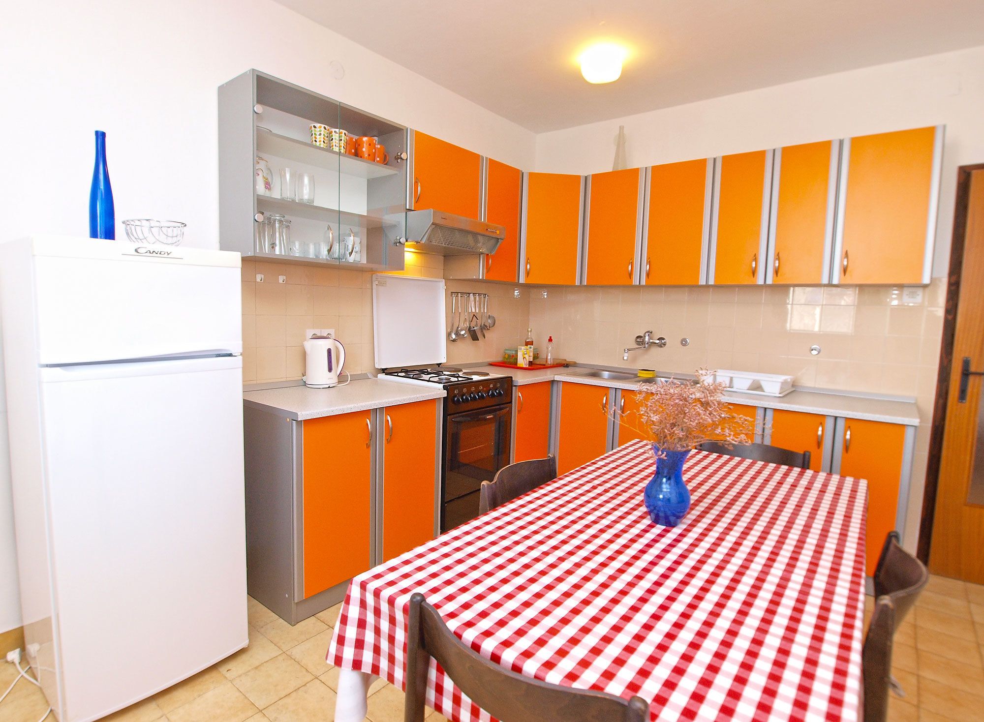 A compact kitchen with orange cabinets, a white refrigerator, and a red and white checkered tablecloth.