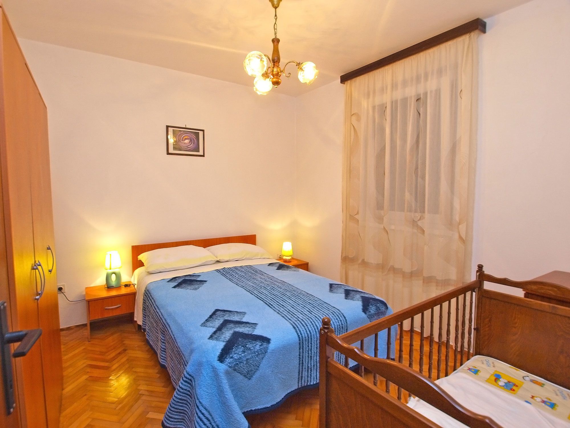 A bedroom with a wooden bed, two bedside lamps, a wooden chair, a wooden floor, a chandelier, and a window with sheer curtains.
