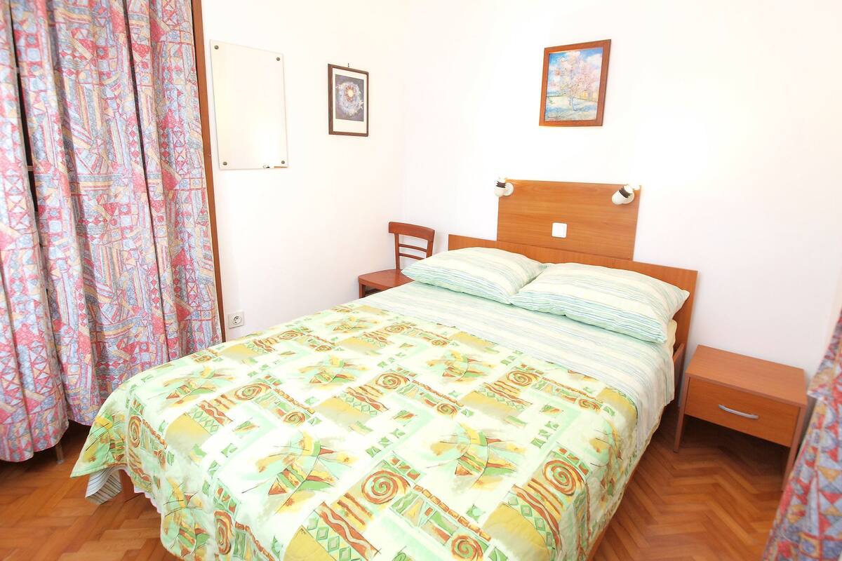 A bedroom with a bed, a nightstand, a wooden headboard, a chair, a curtain, and two framed pictures on the wall.
