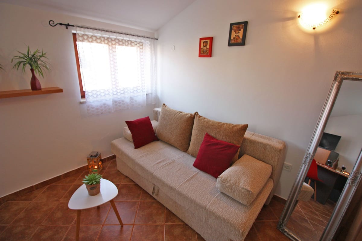 Uma sala de estar com sofá bege, almofadas vermelhas, uma mesa pequena com uma planta, um espelho e uma janela com cortinas leves.