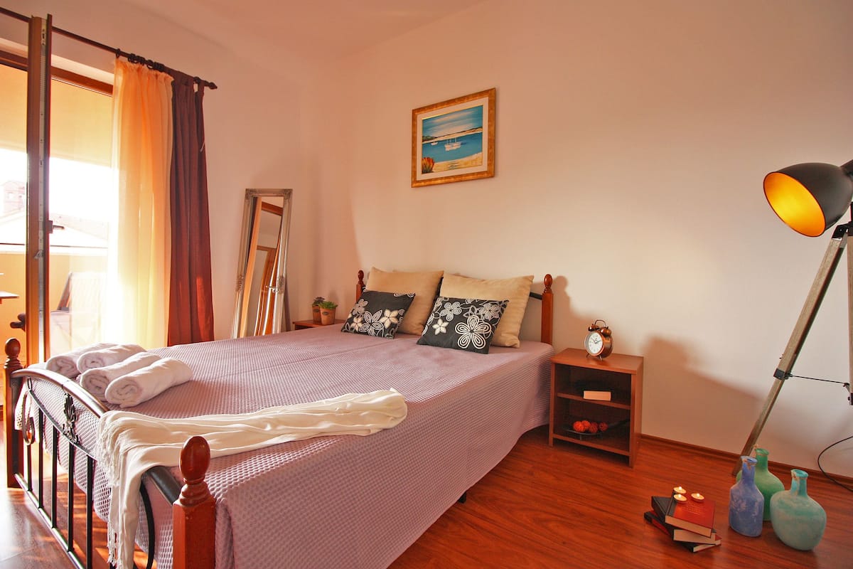 Uma cama bem arrumada com um cobertor branco, uma mesa de cabeceira com abajur e uma pequena estante com livros e objetos decorativos.