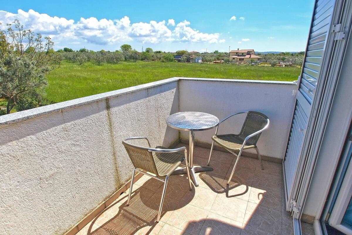 A balcony with a small table and two chairs, overlooking a green field and a building in the distance.