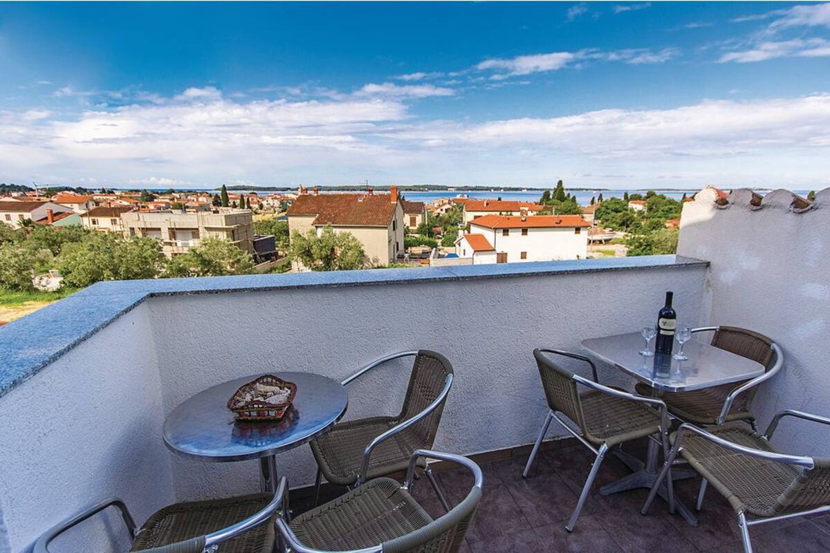 A balcony with wicker chairs and a table, overlooking a coastal town with buildings and a clear sky.