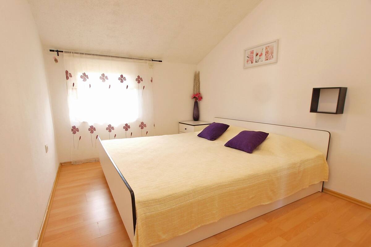 Une chambre à coucher avec un lit, deux oreillers violets, une table de chevet avec un vase et une fenêtre ornée de rideaux à motifs floraux.