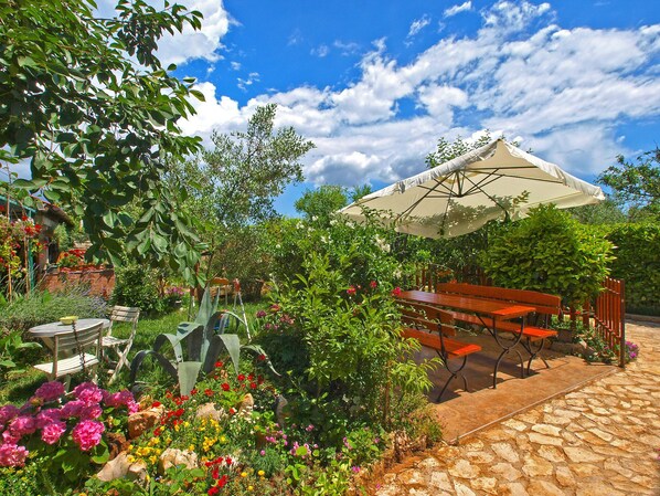 A garden patio with a table and chairs, surrounded by lush greenery and flowering plants.