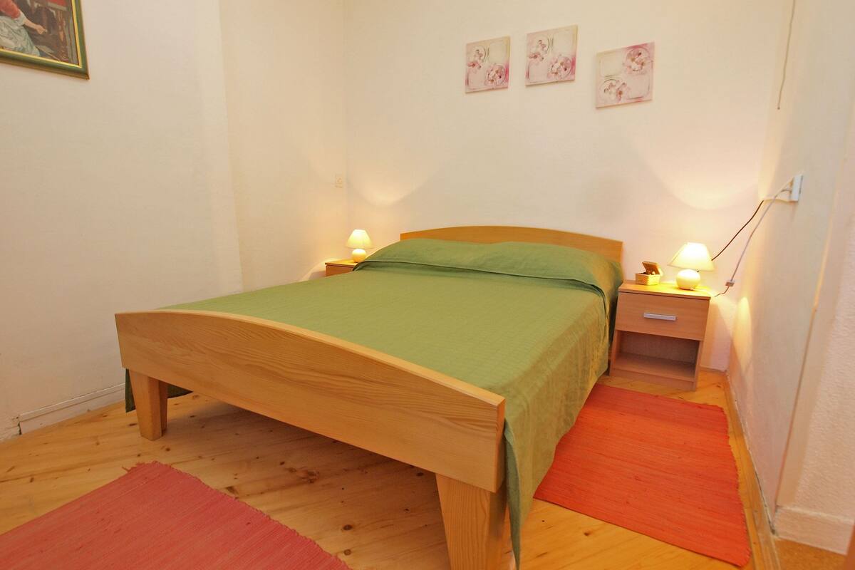 Une chambre avec un lit simple, un cadre en bois, une literie verte, deux tables de chevet avec des lampes et un tapis rouge sur le sol.