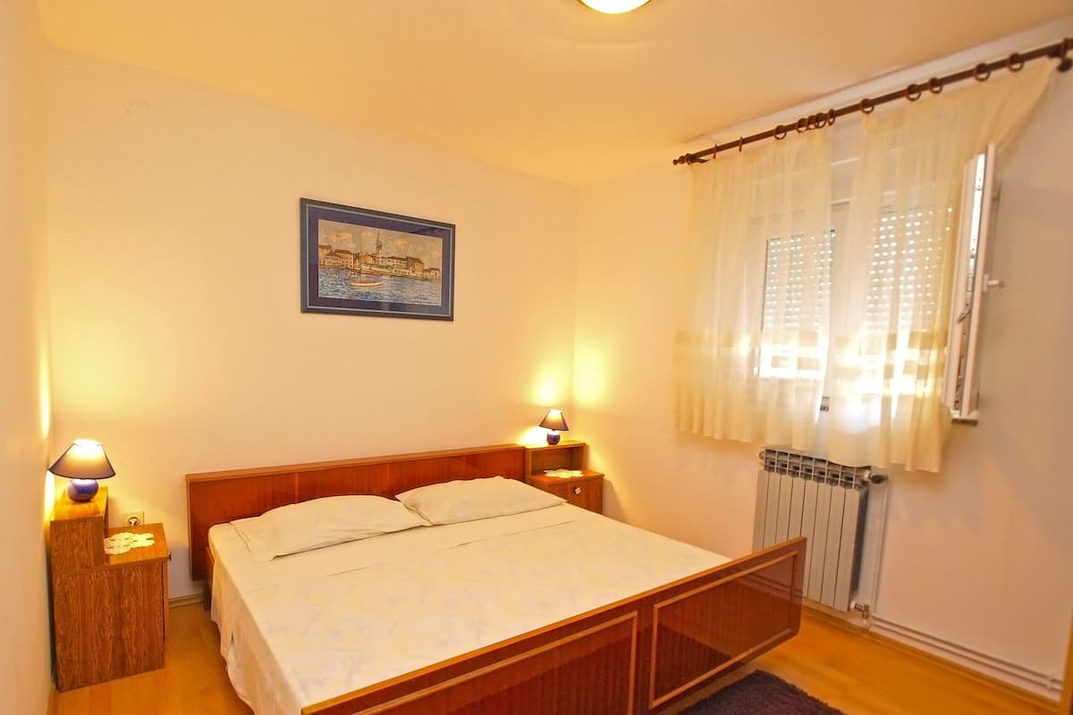 Une chambre à coucher avec un lit en bois, deux lampes de chevet, un tableau encadré au mur, une fenêtre avec des rideaux légers et un radiateur.