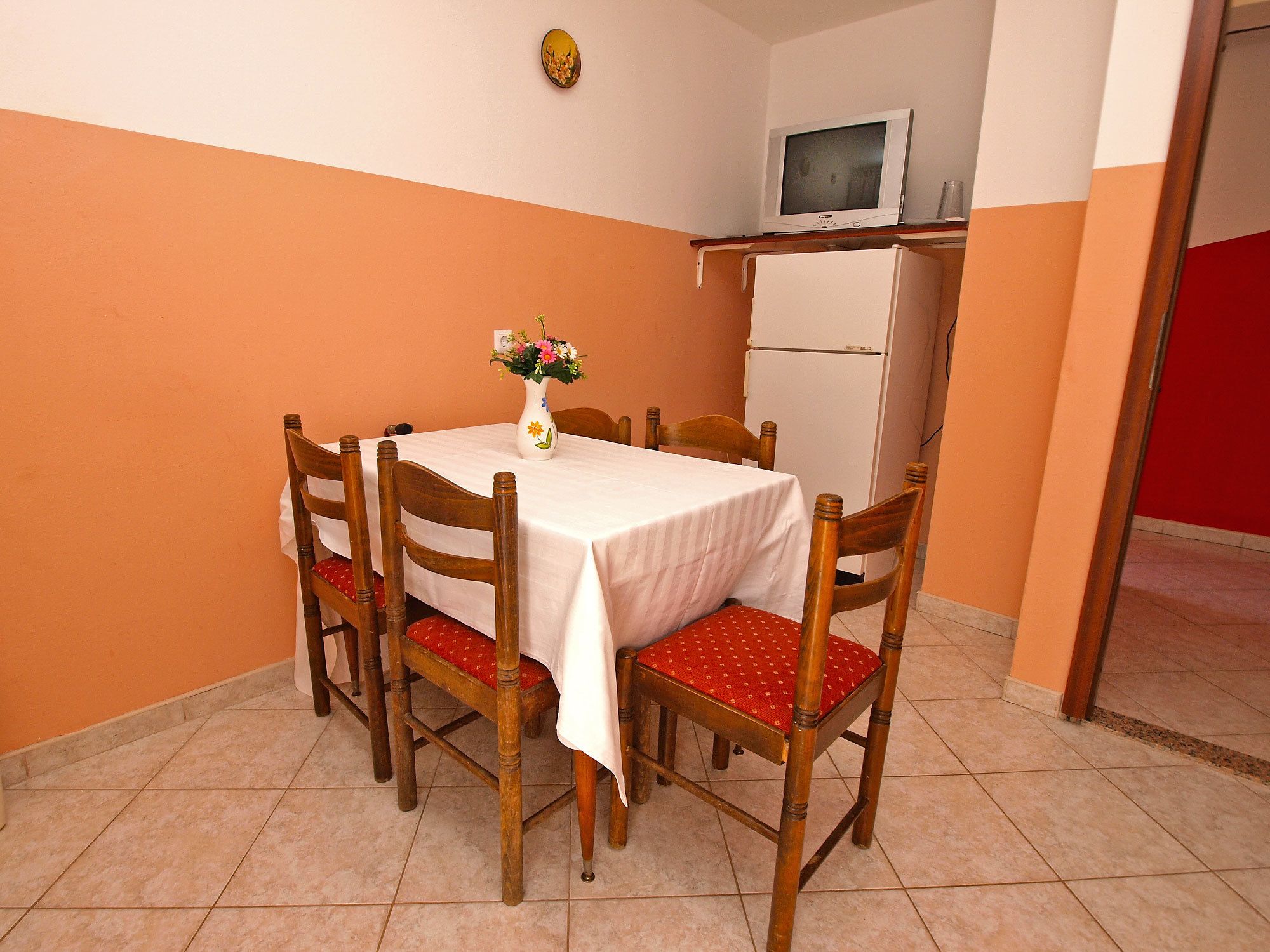 A dining area with a table set for six, surrounded by wooden chairs with red cushions. A white tablecloth covers the table, and a vase with flowers is placed on it. A white refrigerator is visible in the background.