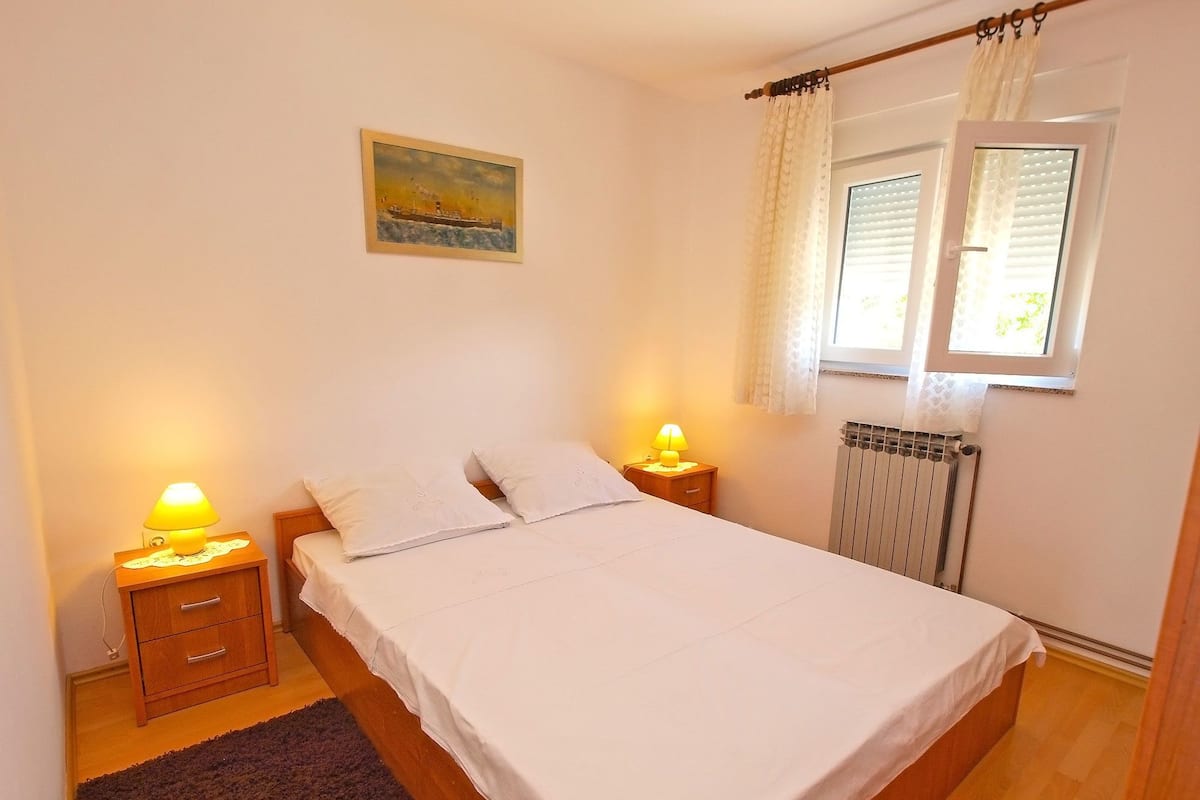 Une chambre à coucher avec un lit, deux tables de chevet avec des lampes, un radiateur, une fenêtre avec des rideaux et un tableau au mur.