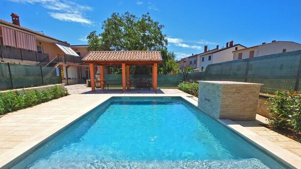 A rectangular swimming pool with clear blue water, surrounded by a tiled deck and a pergola structure. - Apartments Marina 947 (Fazana)