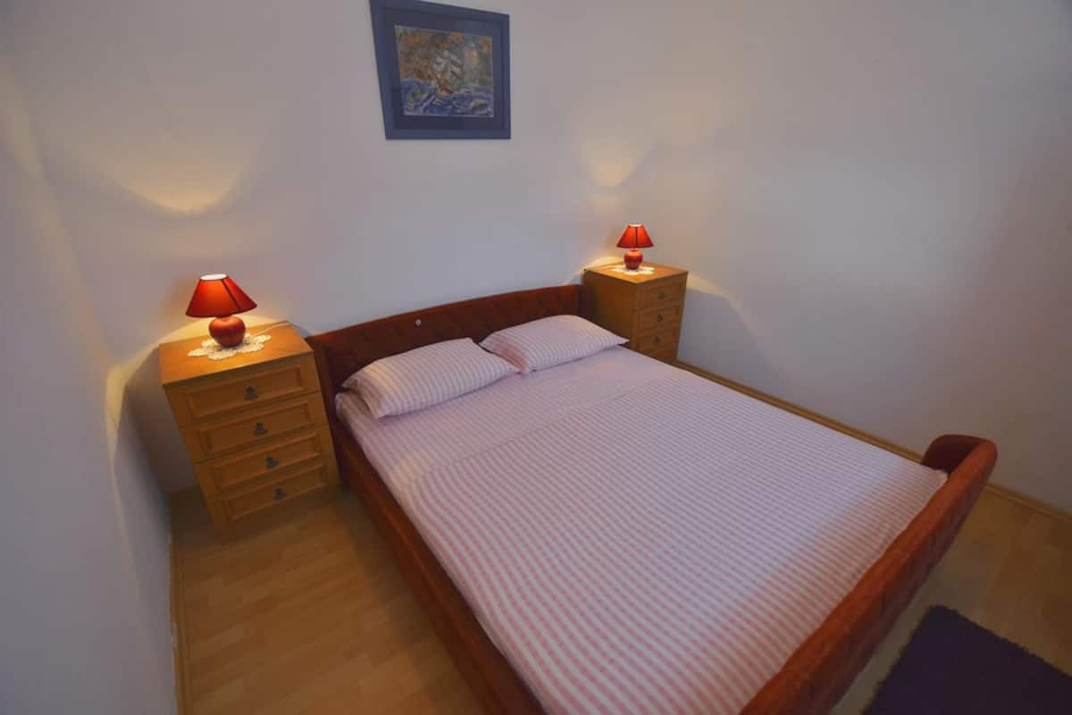 Un lit simple avec une tête de lit en bois, deux tables de chevet avec des lampes, une petite commode et un tableau encadré accroché au mur.