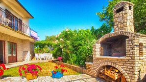 Cocina de piedra al aire libre con chimenea, rodeada por un jardín con plantas florecientes y un patio empedrado.