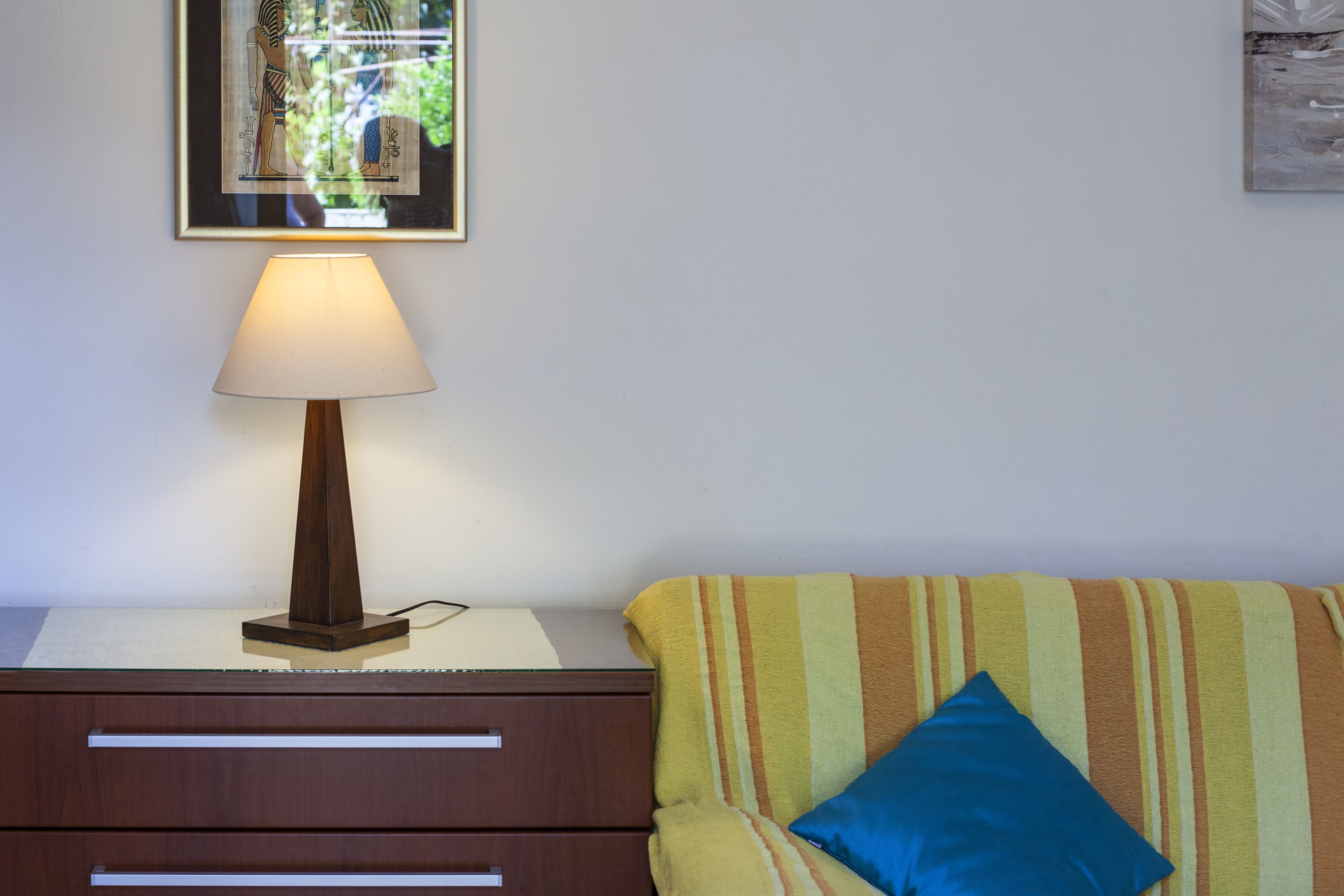 A room with a wooden nightstand, a lamp, a striped sofa, and a framed picture on the wall.