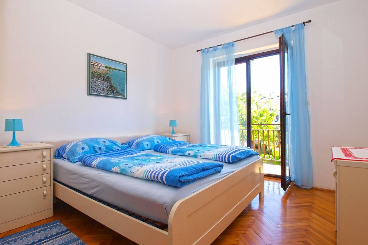 Un dormitorio con cama, mesita de noche, cómoda, ventana con cortinas y balcón.