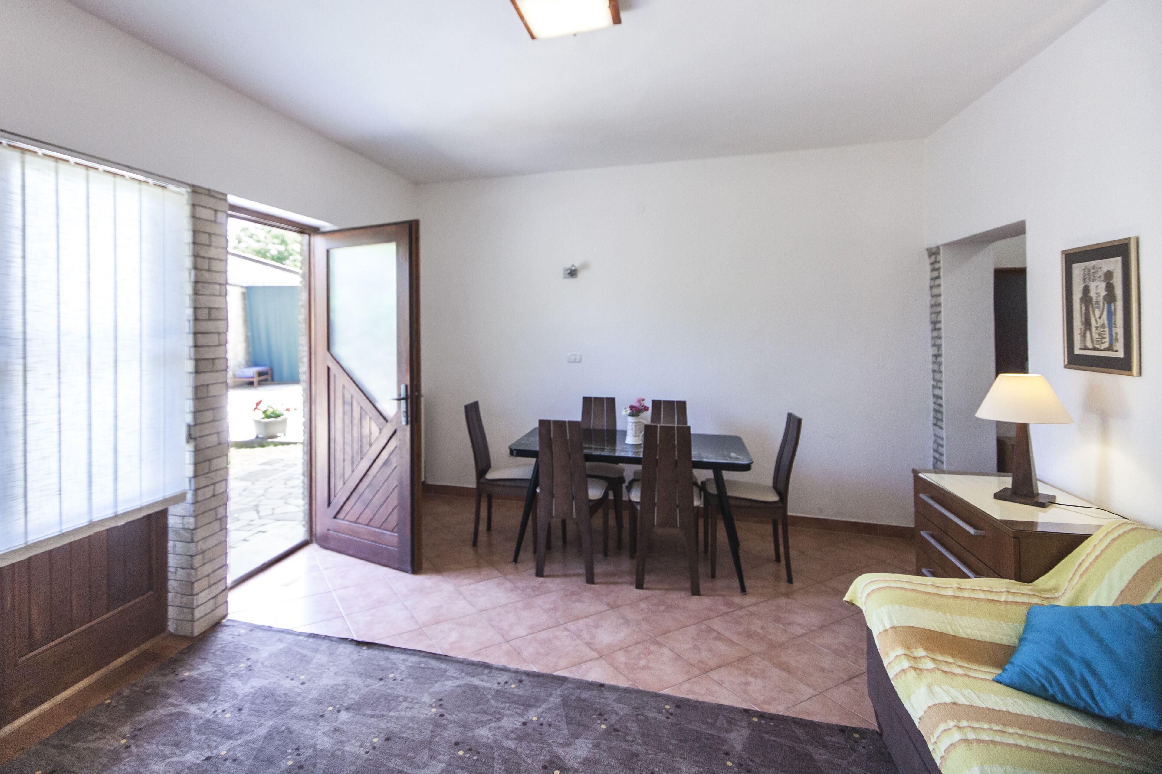 A room with a dining table, chairs, a sofa, a lamp, and a door leading outside.