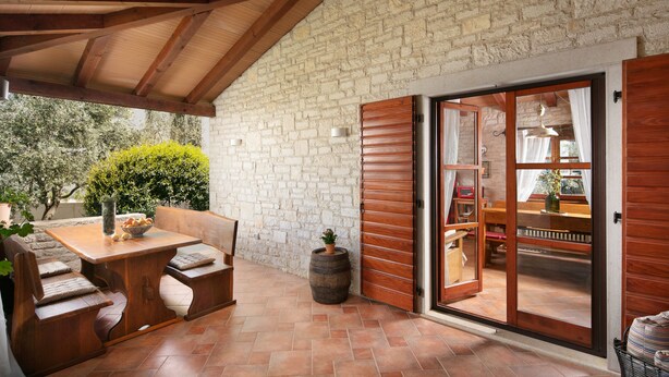 An outdoor patio area with a wooden table and benches, a stone wall, and a sliding glass door leading to an interior space with a bookshelf and a dining table. - Apartments Valter 881 (Novigrad)