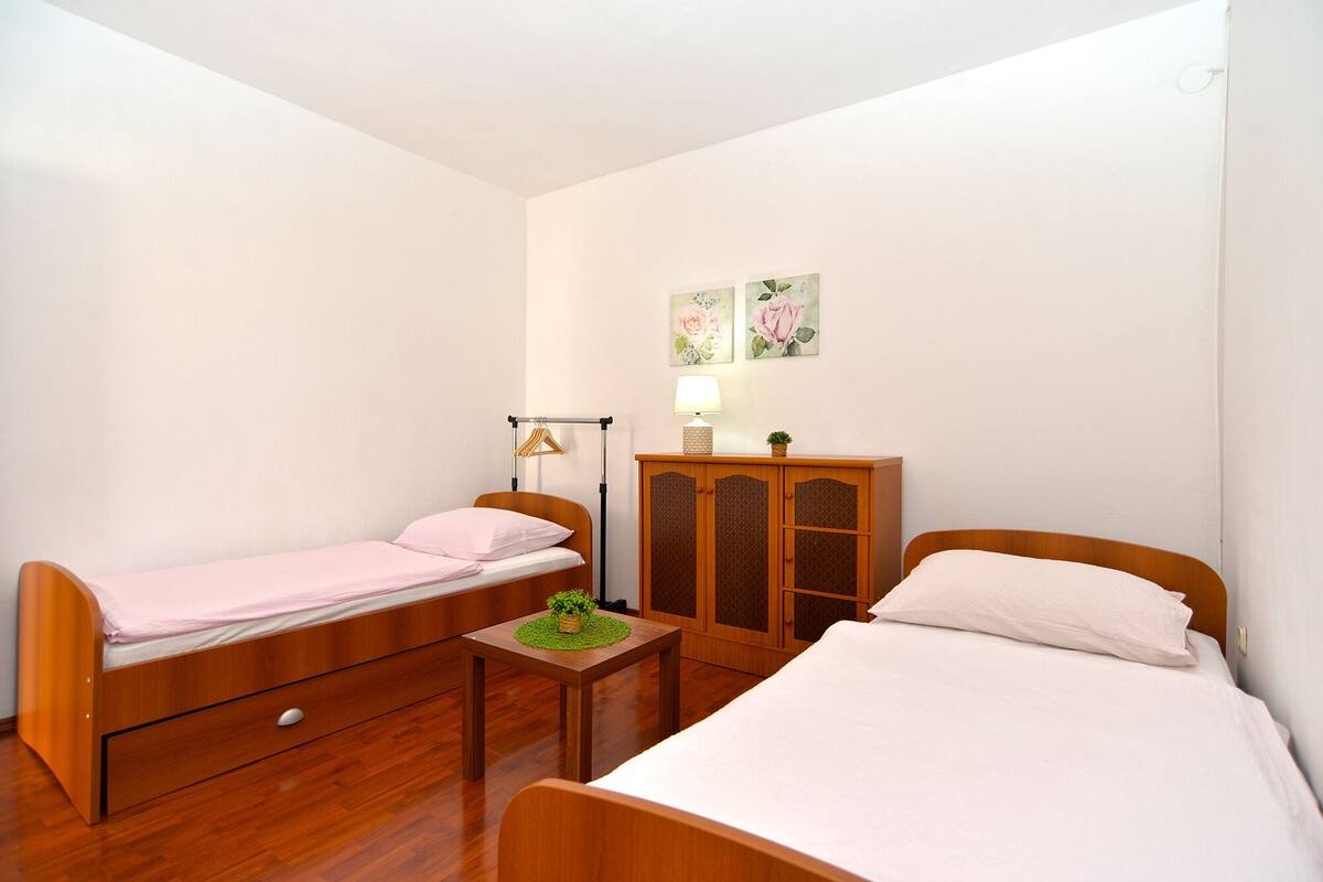 A small, well-lit room with two wooden beds, a wooden cabinet, and a small table with a lamp.