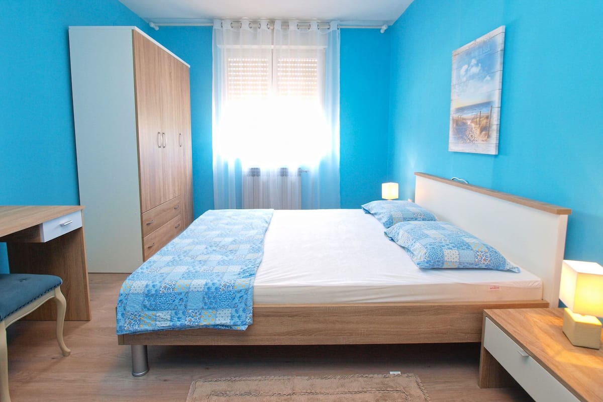 A bedroom with a bed, bedside table, lamp, wardrobe, and a desk with chair.