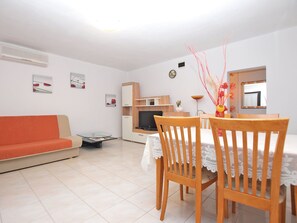 Ein Wohnzimmer mit einer Couch, einem Esstisch mit Stühlen, einem Fernseher und einer kleinen Küchenzeile.