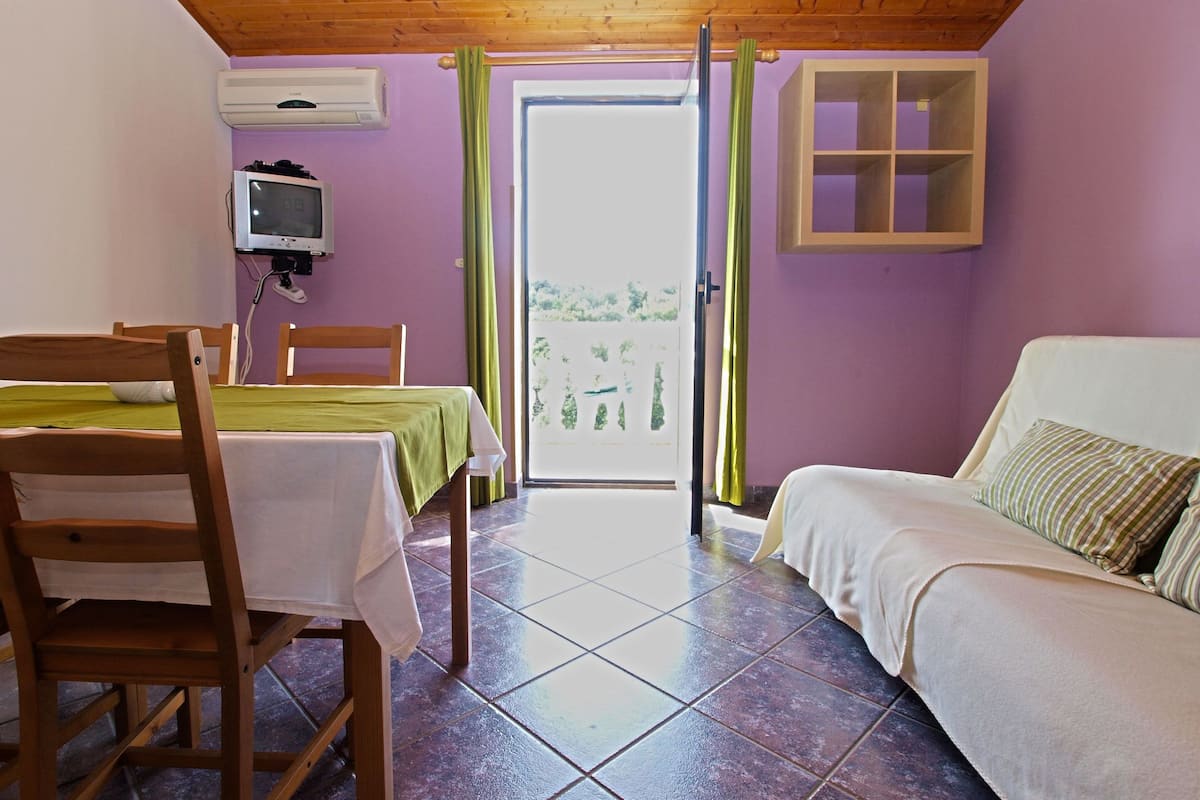 Une pièce avec un lit, une table recouverte d’une nappe verte, une chaise en bois, un téléviseur fixé au mur et une fenêtre avec des rideaux.