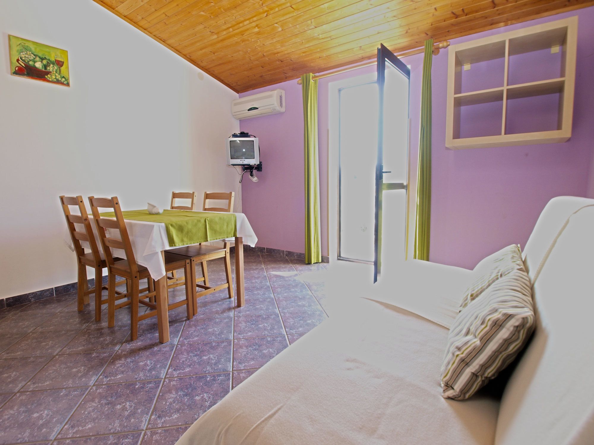 A dining area with a table and chairs, a television mounted on the wall, and a sliding glass door leading to a balcony.