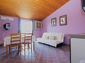 A room with a wooden ceiling, purple walls, a white sofa, a dining table with chairs, a small TV, and framed pictures on the wall.