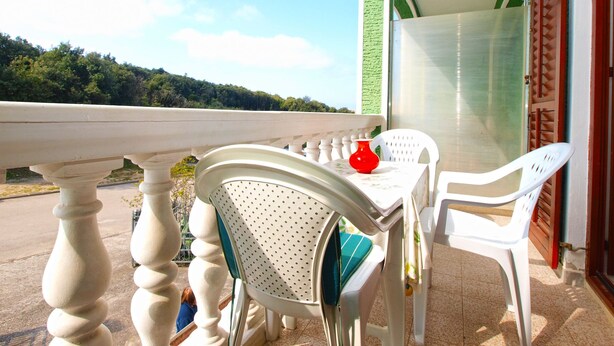 Een balkon met een tafel, stoelen, een rode vaas en uitzicht op een groen landschap.