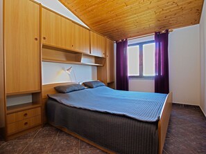 A bedroom with a wooden ceiling, a bed with a grey bedspread, two pillows, a nightstand with a lamp, and a window with purple curtains.