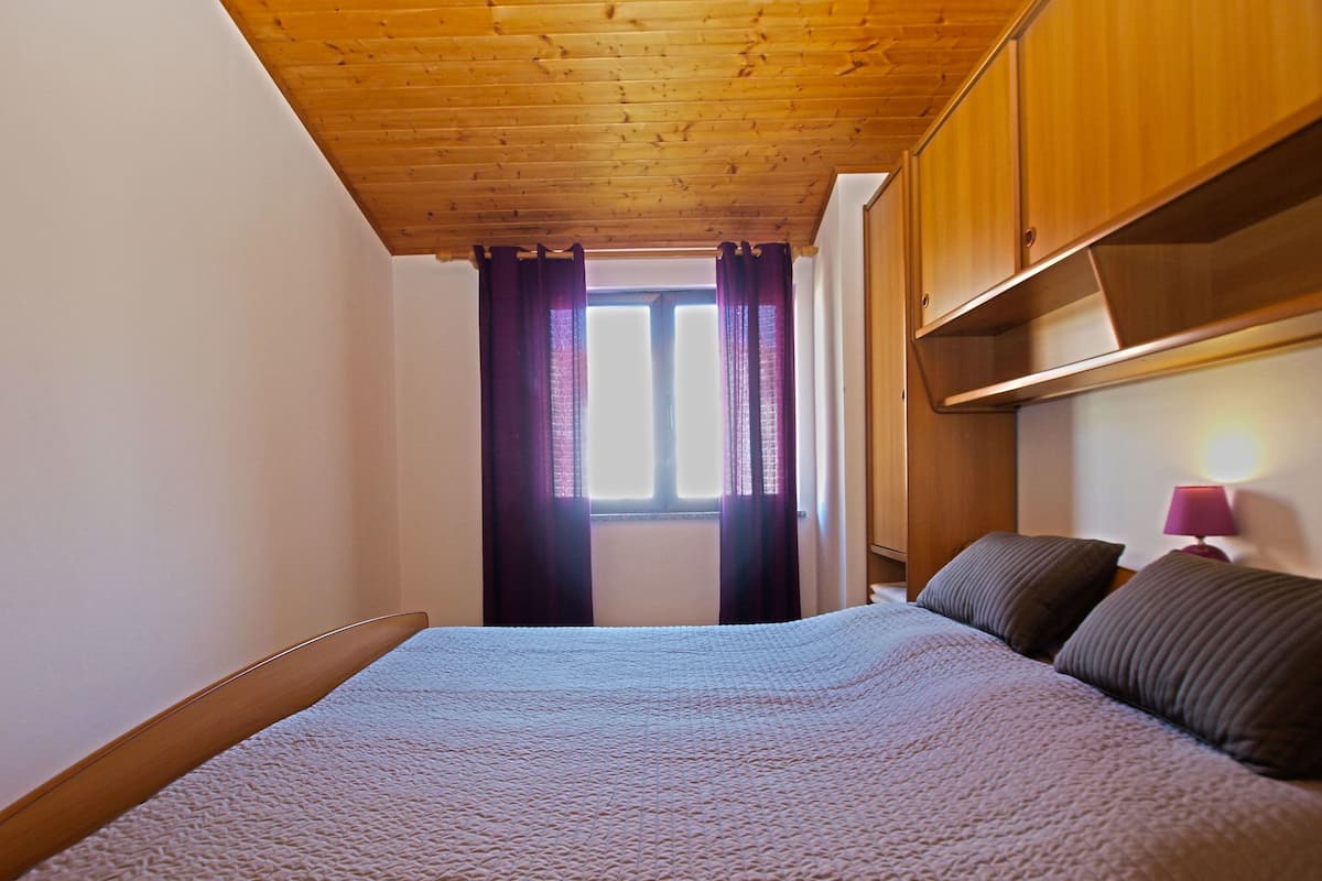 Une chambre avec un plafond en bois, un lit recouvert d’une couverture à motifs, une fenêtre ornée de rideaux violets et une lampe de chevet.