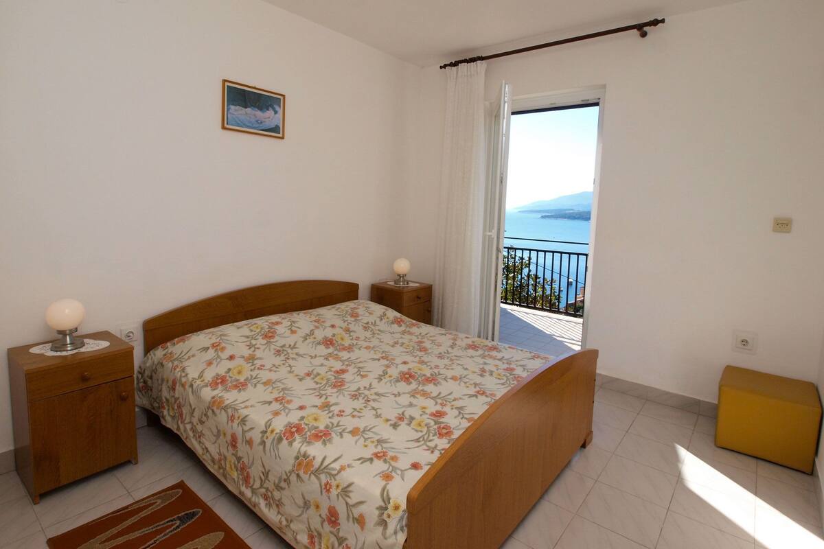 Un dormitorio con cama, mesitas de noche, taburete pequeño, ventana con vista al mar y cortina.