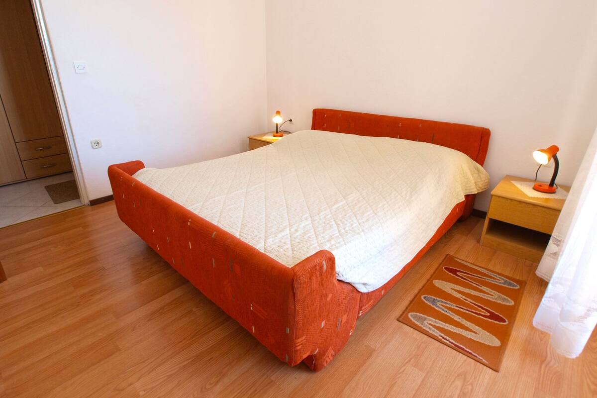 Un dormitorio con cama, dos mesitas de noche con lámparas, suelo de madera y una puerta a la izquierda.