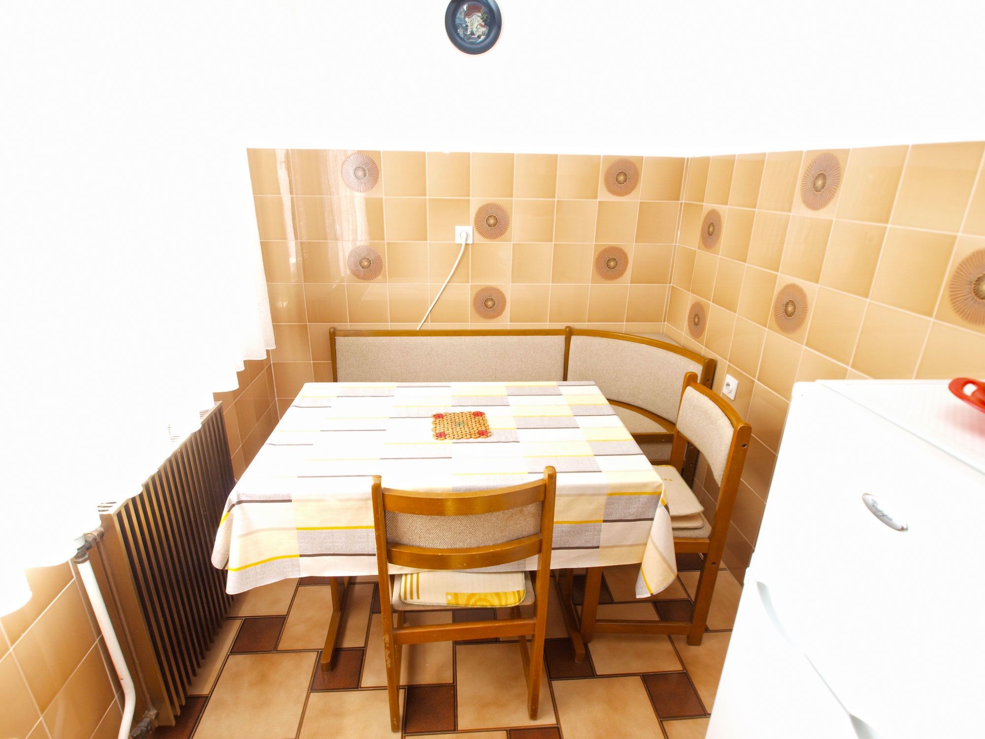 Een kleine keuken met een betegelde spatwand, een centrale tafel met stoelen, en een witte koelkast.