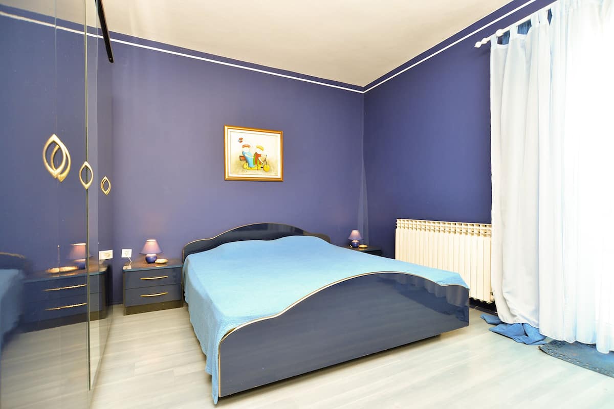 Quarto com cama, mesas de cabeceira, abajur, pintura e janela com cortinas.