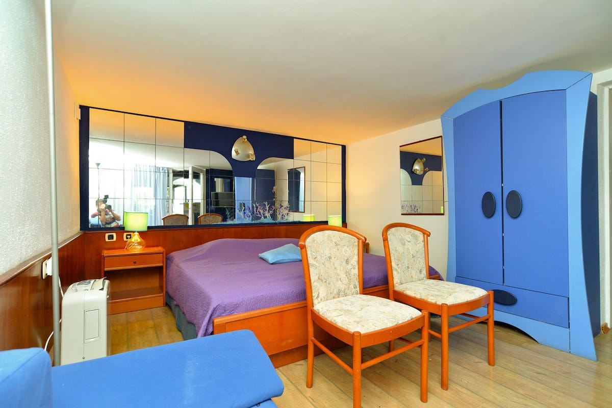 Um quarto com cama, duas cadeiras, guarda-roupa, espelho e mesa de cabeceira.