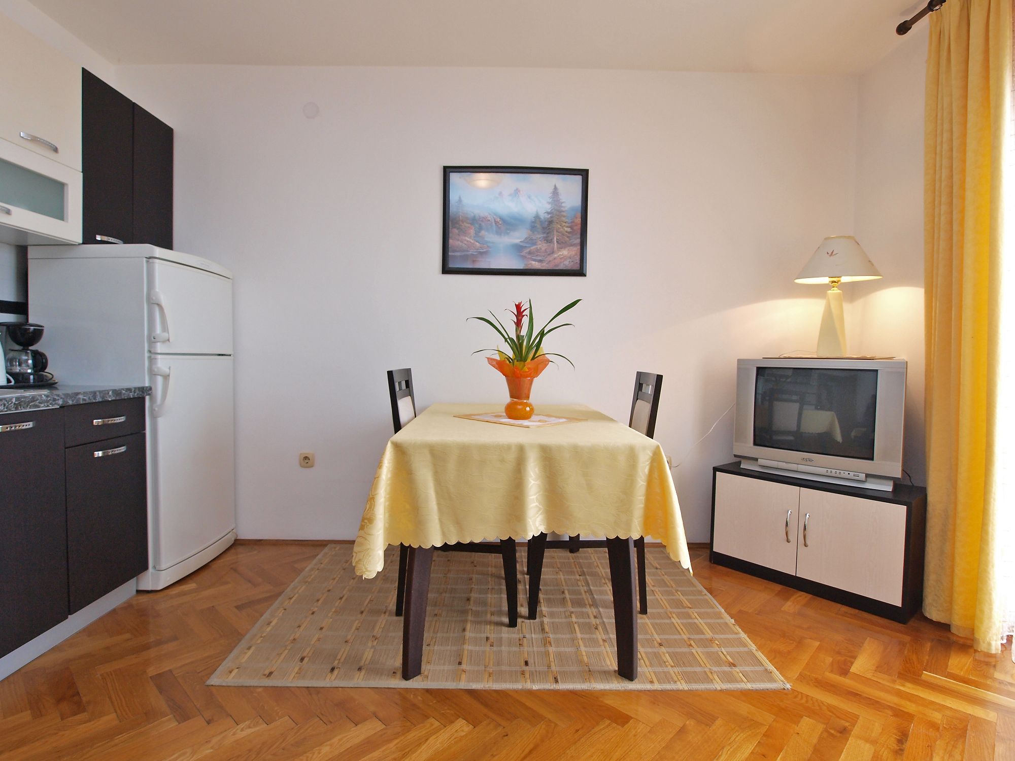 A compact living space with a dining table, chairs, a small kitchen area, a television, and a framed picture on the wall.