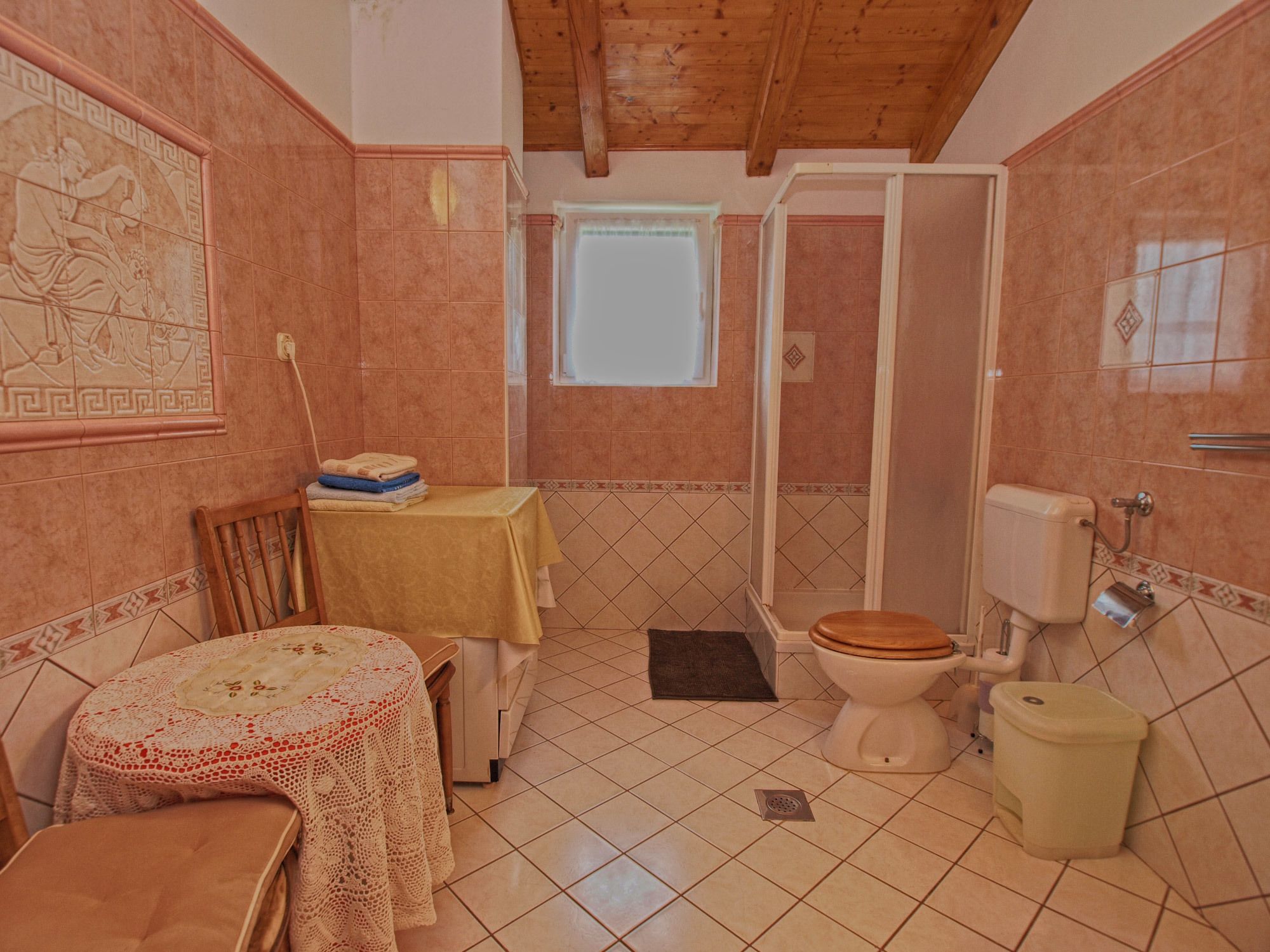 A bathroom with a toilet, sink, and shower. There is a table with a towel and a chair.