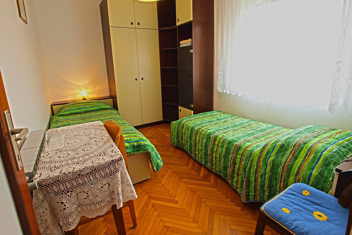 Une petite chambre à coucher avec deux lits simples, un plancher en bois, une table recouverte d’une nappe en dentelle et une armoire.
