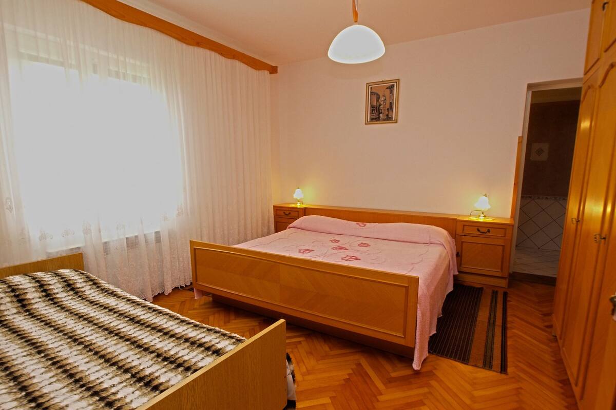 Une chambre à coucher avec un lit en bois, une table de chevet avec une lampe, une fenêtre avec des rideaux légers et une porte donnant sur une autre pièce.