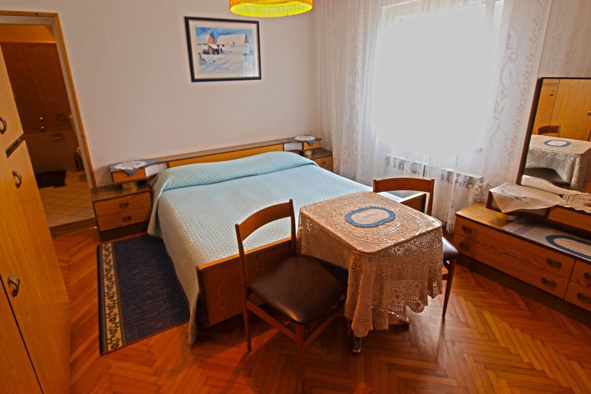 Une chambre à coucher avec un lit, un bureau en bois, une chaise, une table recouverte d’une nappe en dentelle, une lampe suspendue, un tableau encadré au mur et une fenêtre avec des rideaux.