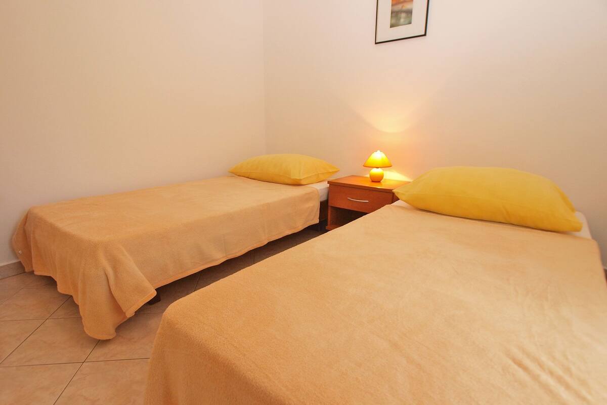 A room with two beds, each with a yellow pillow and a matching blanket. There is a nightstand with a lamp between the beds.