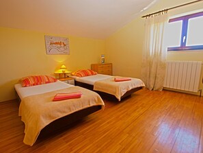 Una habitació amb dos llits, terres de fusta, un radiador i una finestra amb cortines.