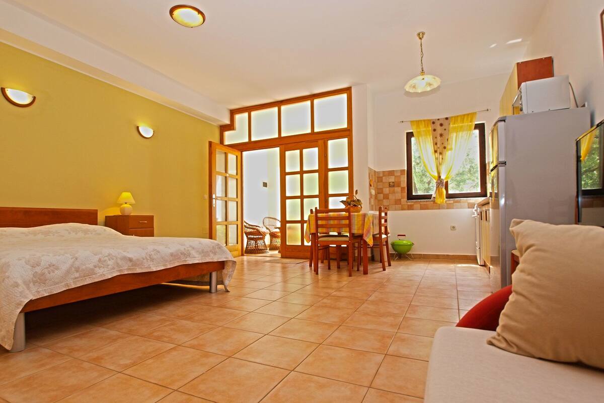 A bedroom with a bed, a nightstand, a table with chairs, and a kitchenette.