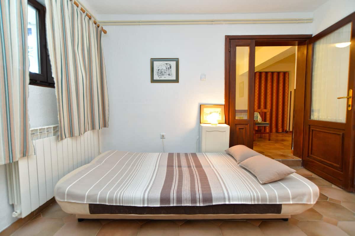 A bedroom with a bed, a bedside lamp, a framed picture on the wall, and a door leading to another room.