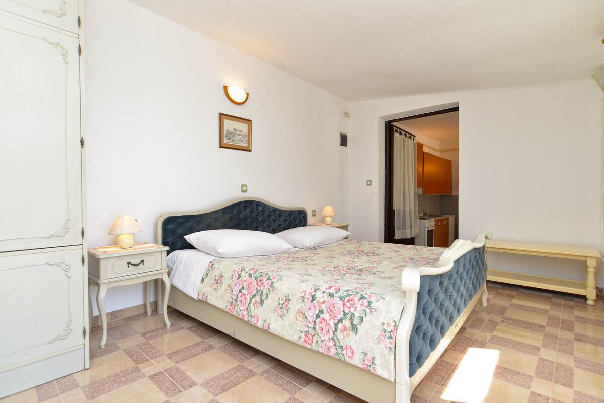 A bedroom with a bed, bedside tables, a lamp, a framed picture on the wall, and a view into the kitchen.