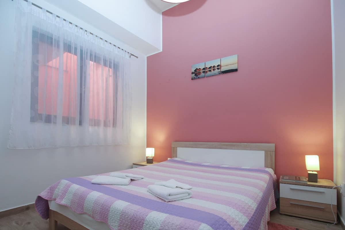 Ein Schlafzimmer mit einem Bett, zwei Nachttischen mit Lampen, einem Fenster mit durchscheinenden Vorhängen und einem an der Wand hängenden Bild.