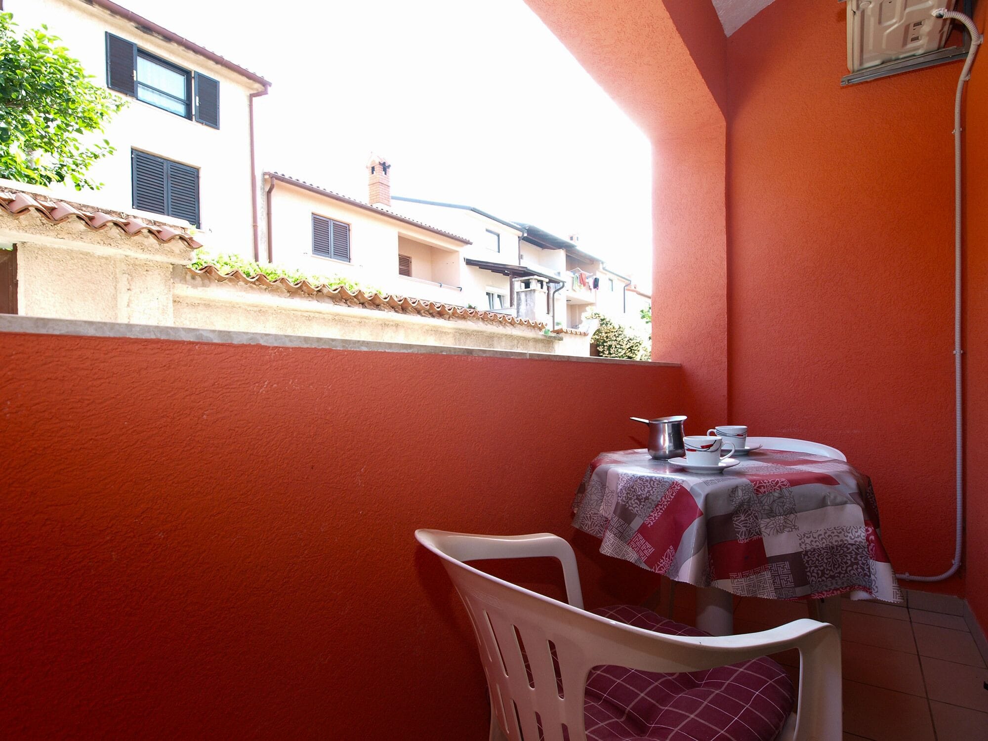 Un balcone con un tavolo apparecchiato per due, una sedia di plastica bianca e un muro rosso.